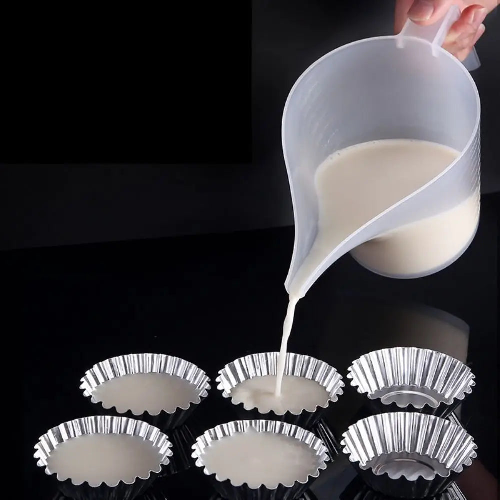 A hand pours white liquid from a plastic measuring cup with a long spout into small metal cups.