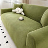 A textured olive green stretch jacquard sofa cover with a white decorative pillow and a cup on the cushion.