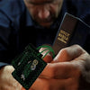 A hand holds a green scapular necklace with Saint Jude and Our Lady of Guadalupe against a dark background.