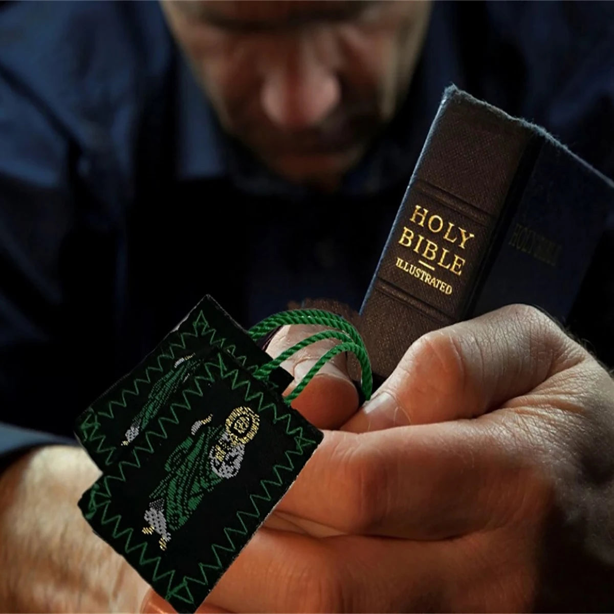 A hand holds a green scapular necklace with Saint Jude and Our Lady of Guadalupe against a dark background.