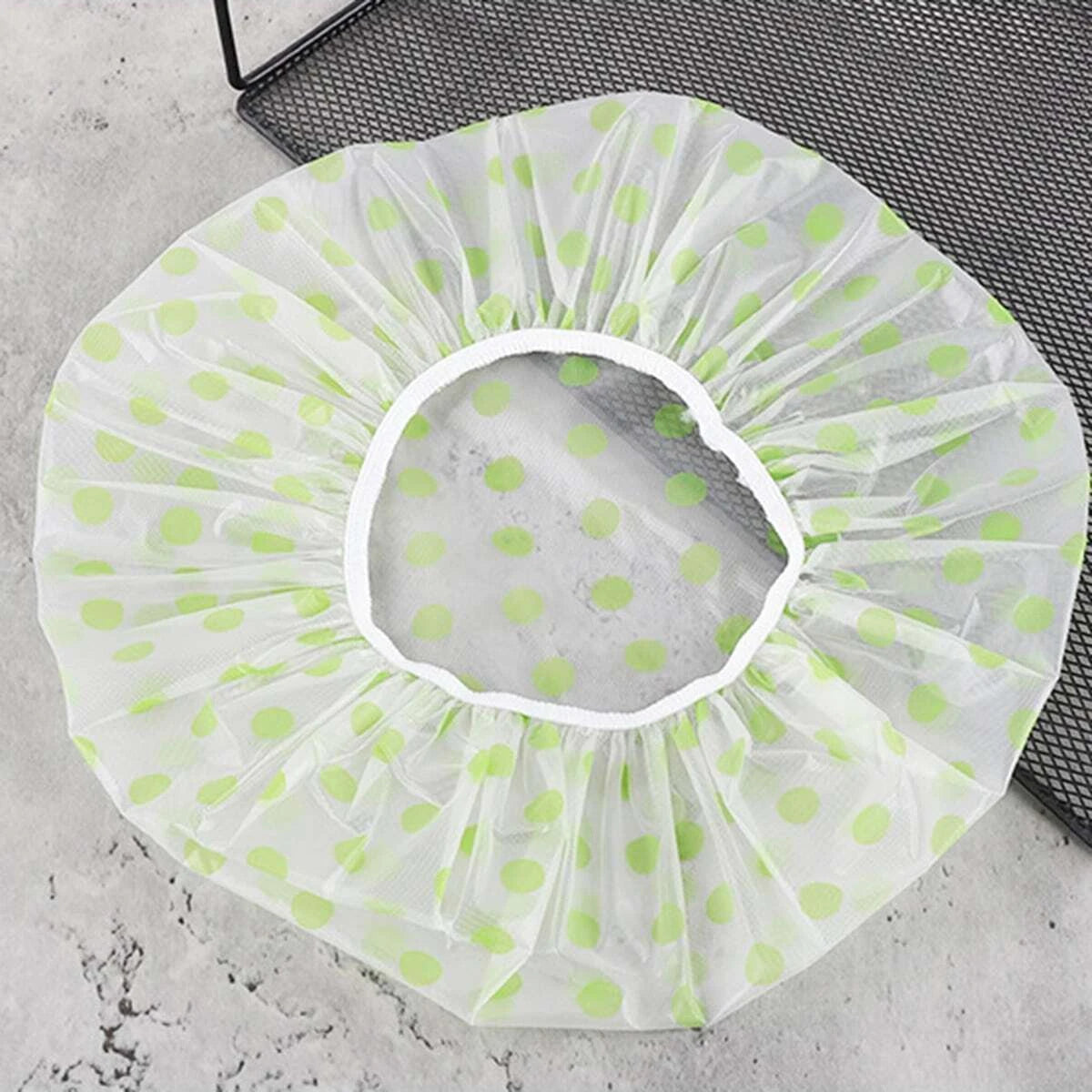 A white disposable waterproof shower cap with light green polka dots is shown on a dark gray surface.