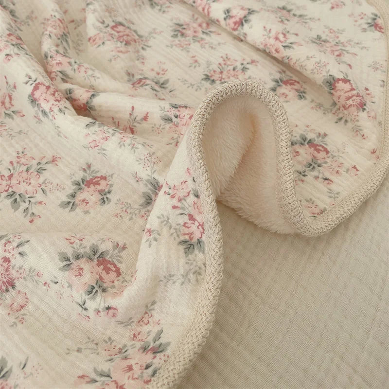 A soft, light pink floral muslin baby blanket with a textured edge is draped and folded.