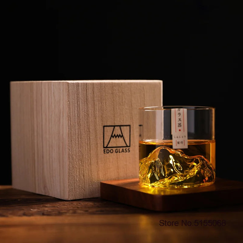 A wooden box holds a glass of amber whiskey with an ice cube on a dark wooden surface.