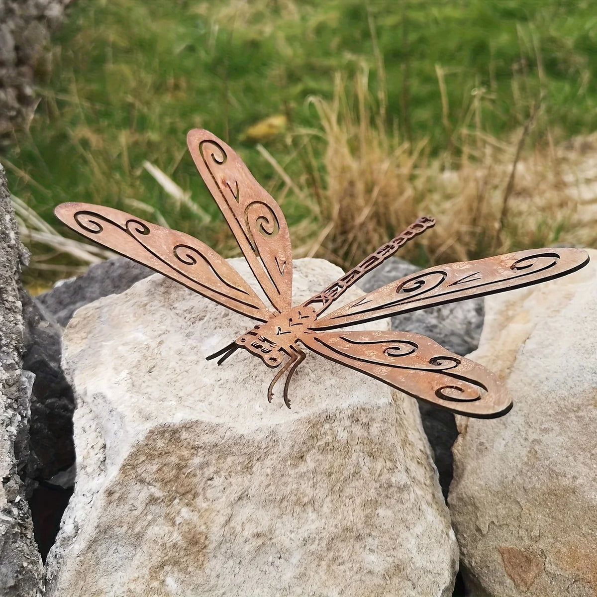 Statue de libellule en métal couleur rouille dans un jardin, posée sur une grande roche claire.