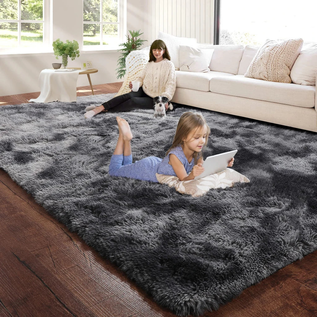 A young girl lies on a dark gray plush carpet in a bright living room with a woman and a dog nearby.
