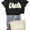 Black t-shirt with a gold glitter cheer letter patch on the chest and a beige canvas tote bag.