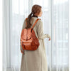 A woman wearing a tan coat is standing indoors, holding a brown vintage faux leather women's backpack.