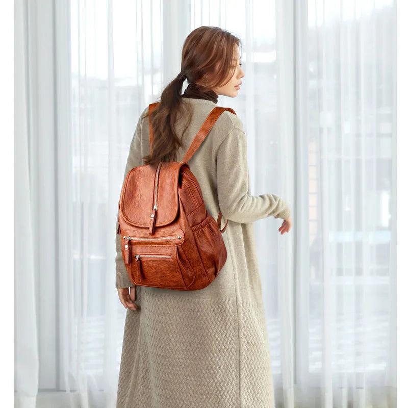 A woman wearing a tan coat is standing indoors, holding a brown vintage faux leather women's backpack.