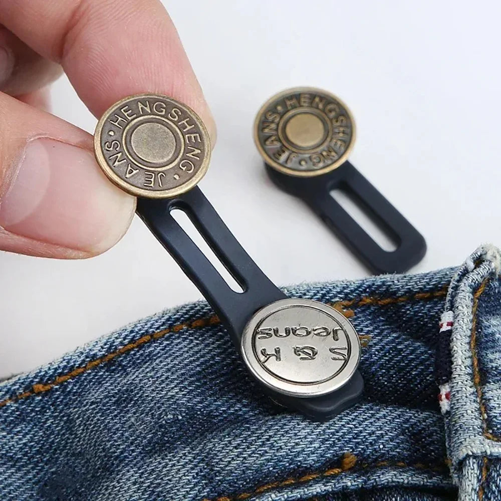 Une main tient un rallonge de taille réglable en laiton pour jeans, montrant les composants métalliques et en plastique noir.