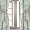 A pair of light green sheer curtains with floral patterns tied back by natural fiber tassels.