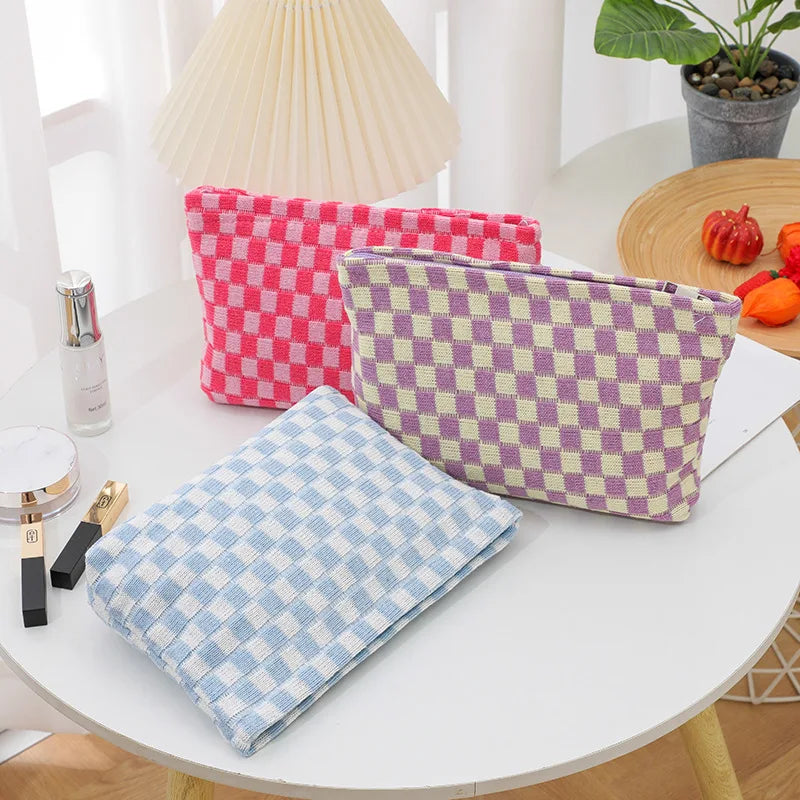 A collection of plaid makeup bags in pink, purple, and blue are displayed on a white table next to a lamp.