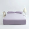 A lavender cooling ice-silk fitted sheet is displayed on a white surface with two pillows and a white lamp.