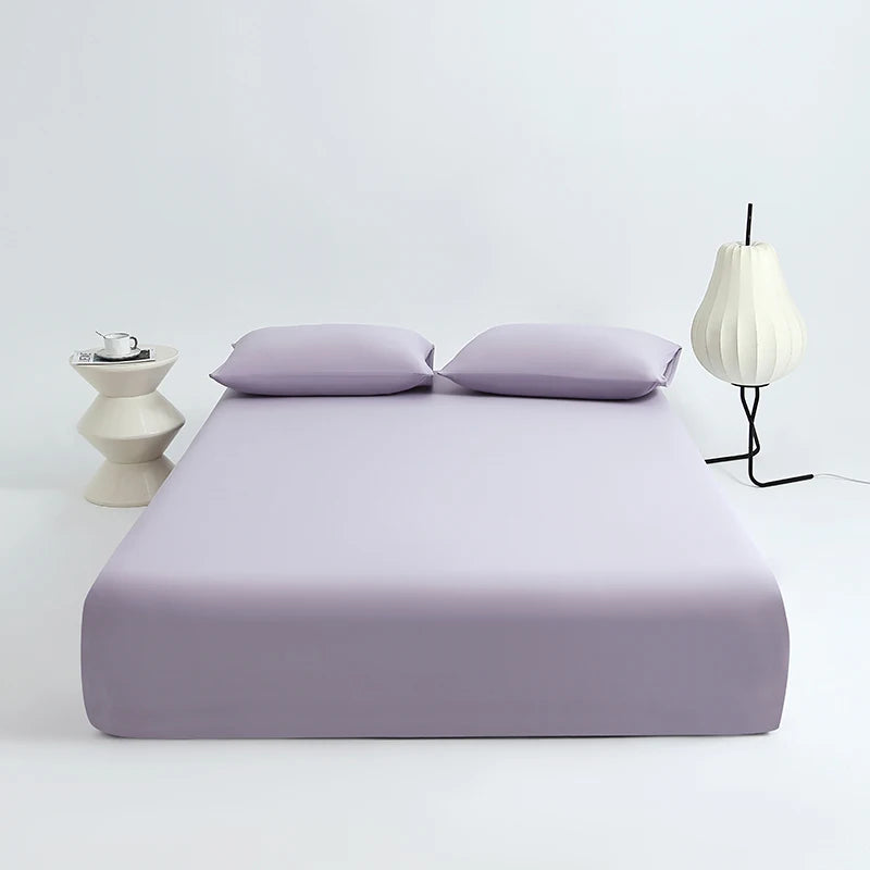 A lavender cooling ice-silk fitted sheet is displayed on a white surface with two pillows and a white lamp.