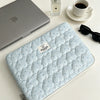 A light blue quilted laptop sleeve with a zipper is shown next to a silver perfume bottle and a white cup on a white surface.