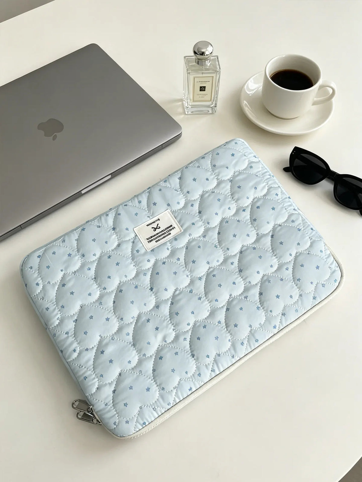 A light blue quilted laptop sleeve with a zipper is shown next to a silver perfume bottle and a white cup on a white surface.