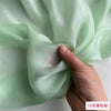 A hand is folding a piece of light green, sheer silk chiffon fabric.