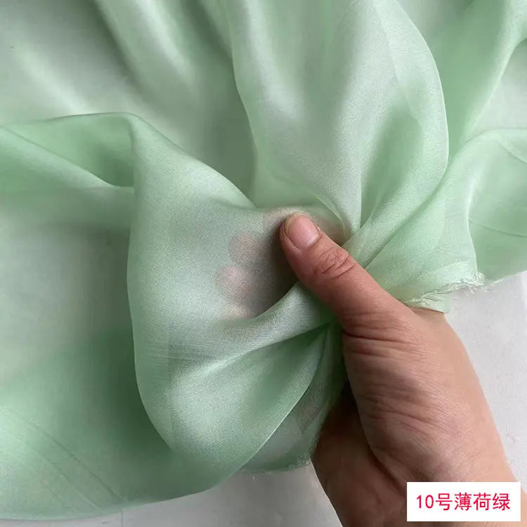 A hand is folding a piece of light green, sheer silk chiffon fabric.