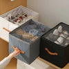 A hand is placing a dark gray foldable shoe organizer with a dustproof lid onto a wooden surface.