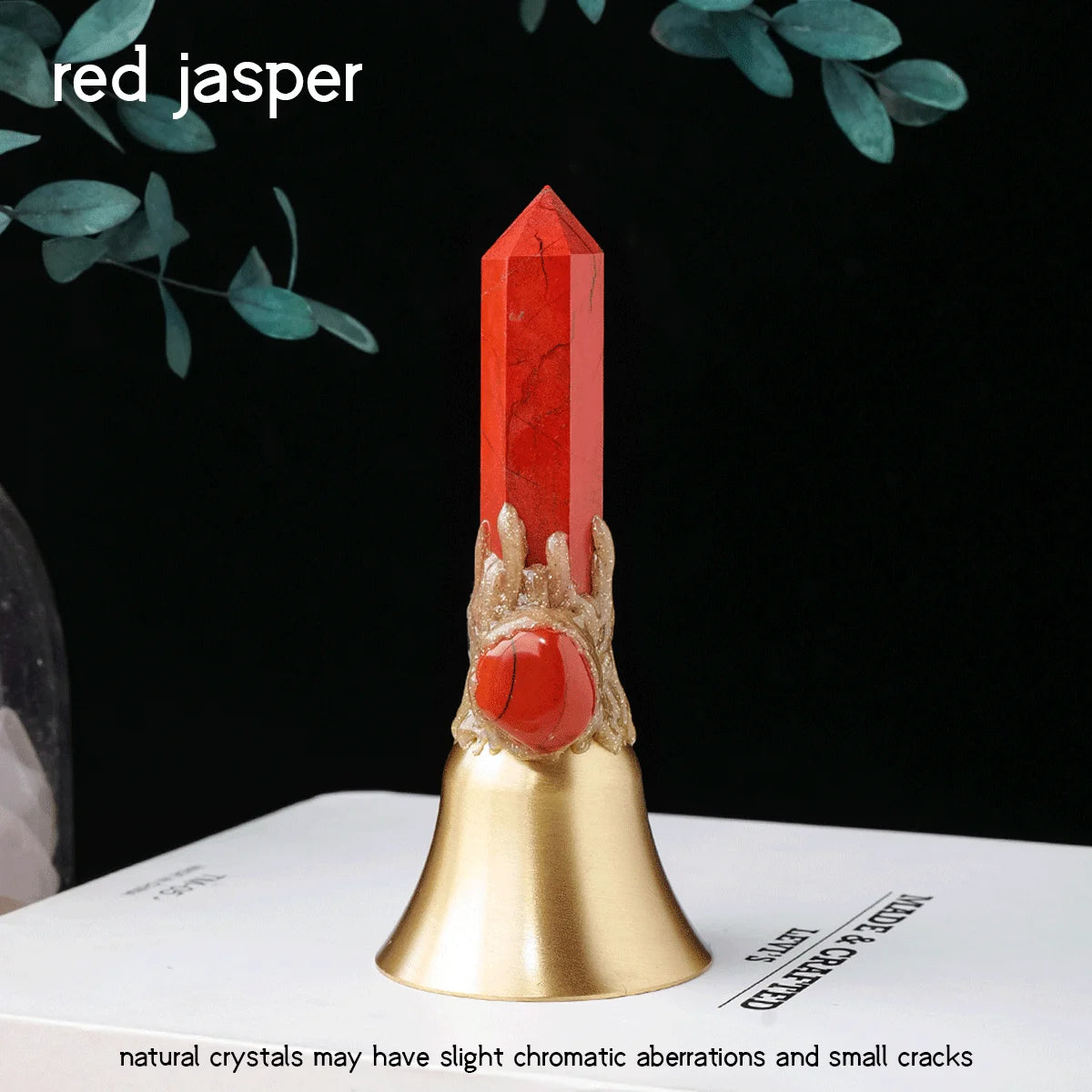 A red jasper crystal point with a brass bell resting on a white surface.