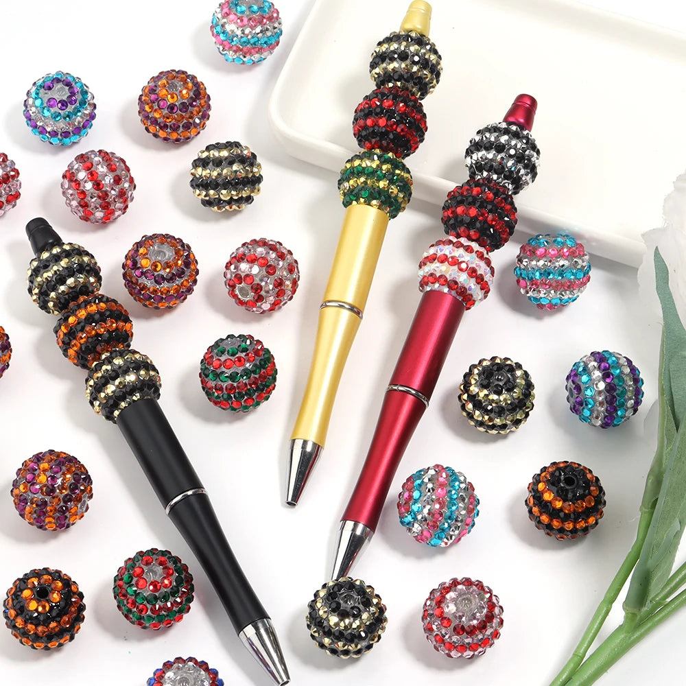 A set of five resin rhinestone beads with a two-tone striped design are displayed on a white tray alongside a pen.