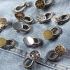 A collection of silver and brass adjustable elastic waist extender buttons laid out on a denim fabric.
