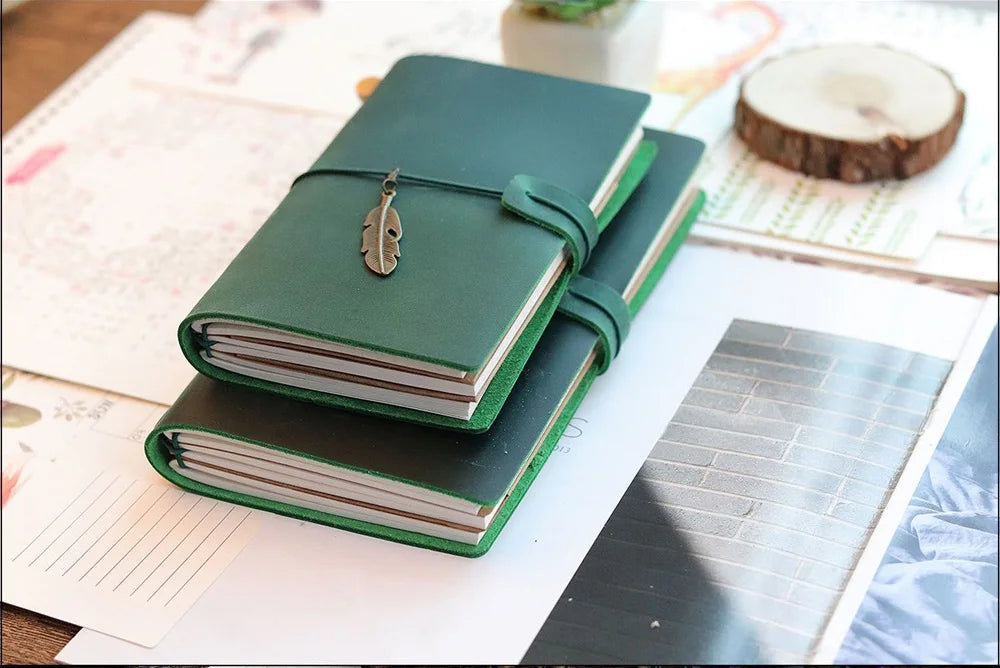 A dark green handcrafted leather refillable journal lying on a light surface next to paper and a wooden slice.