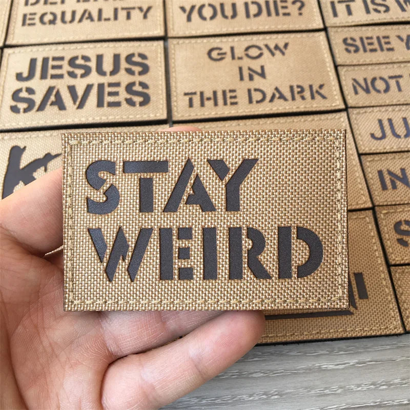 A hand holds an IR reflective Stay Weird tactical patch with hook-and-loop backing against a background of similar patches.