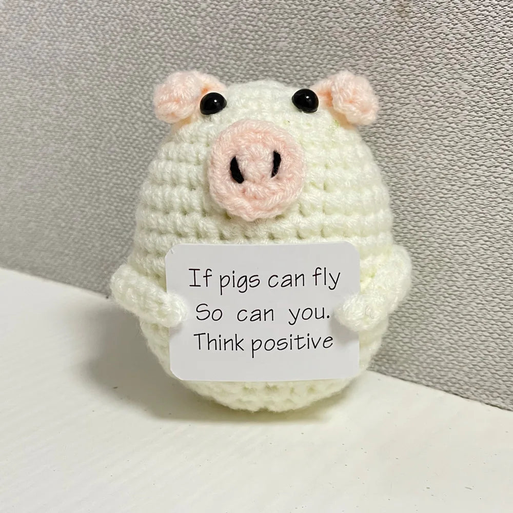 A handmade crochet doll shaped like a pig with pink snout and black eyes, featuring text encouraging positivity.
