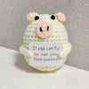 A handmade crochet doll shaped like a pig with pink snout and black eyes, featuring text encouraging positivity.