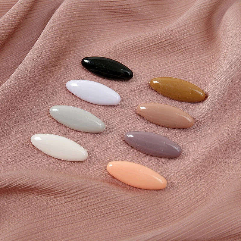 A set of eight solid color scarf pins in black, yellow, white, and pink shades arranged on a pink fabric.