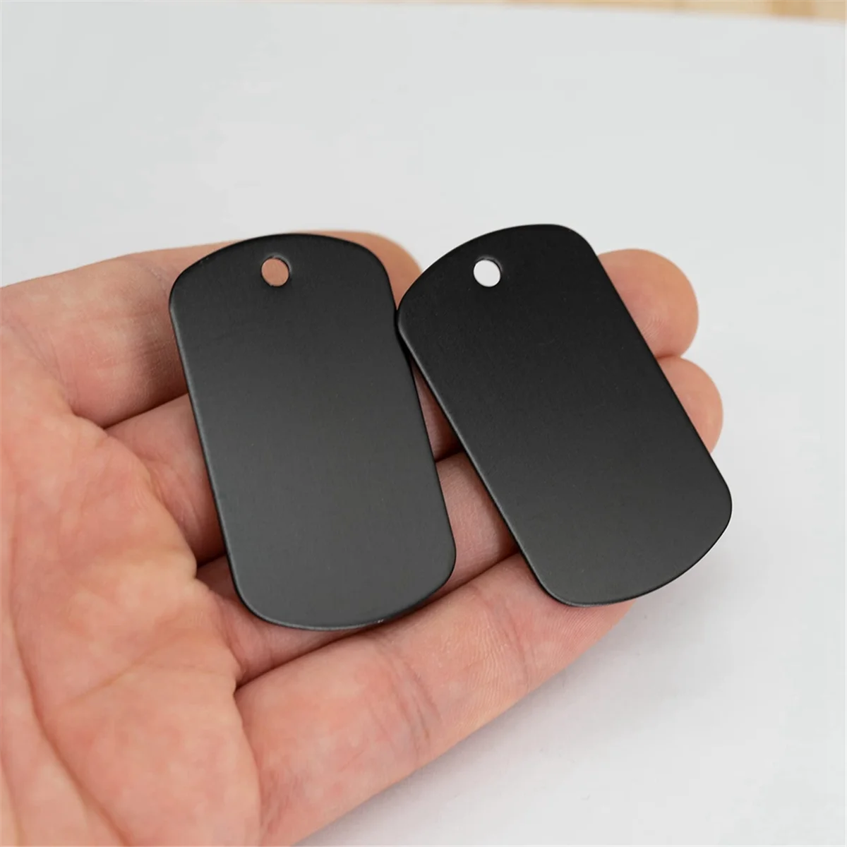 Two black aluminum dog tags are held in a person's hand against a white background.