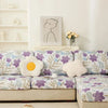 A floral elastic sofa cover in purple and green patterns on a beige couch with a white wall and framed artwork.