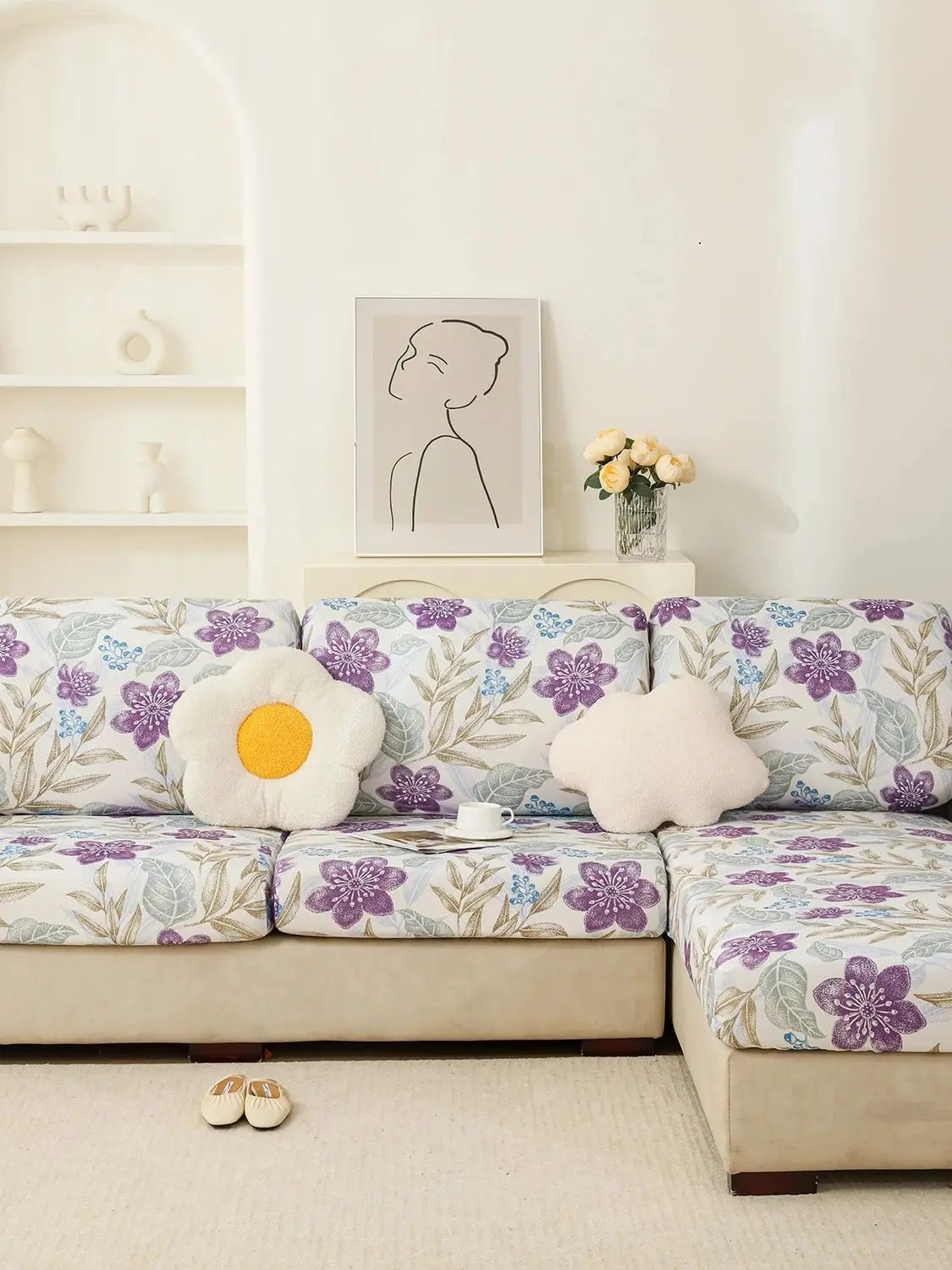 A floral elastic sofa cover in purple and green patterns on a beige couch with a white wall and framed artwork.
