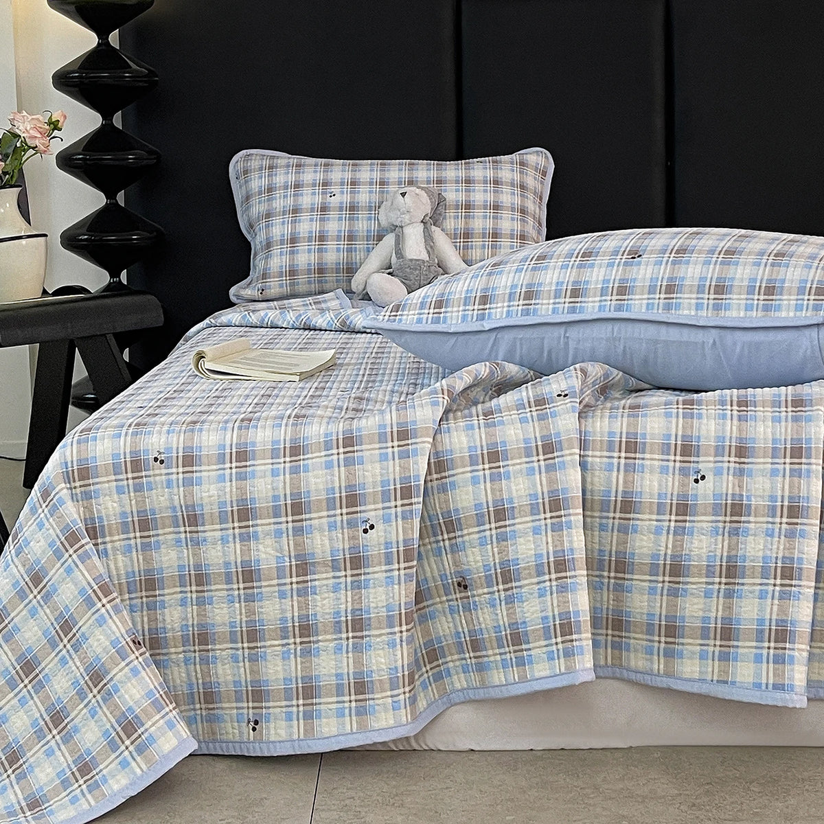 A plaid blue and beige quilted bedspread with a stuffed animal pillow on a dark background.