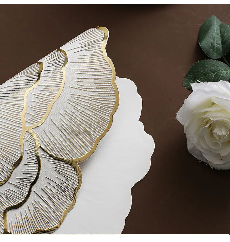 Goldvergoldete Pfingstrosen-Platzsets mit gerippter Struktur auf einer dunkelbraunen Oberfläche neben einer weißen Rose.