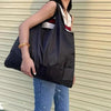A person is holding a black foldable nylon shopping tote bag with a white and red accent on their shoulder.