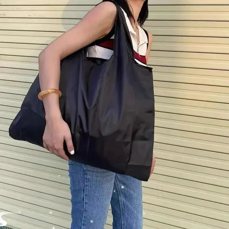 A person is holding a black foldable nylon shopping tote bag with a white and red accent on their shoulder.