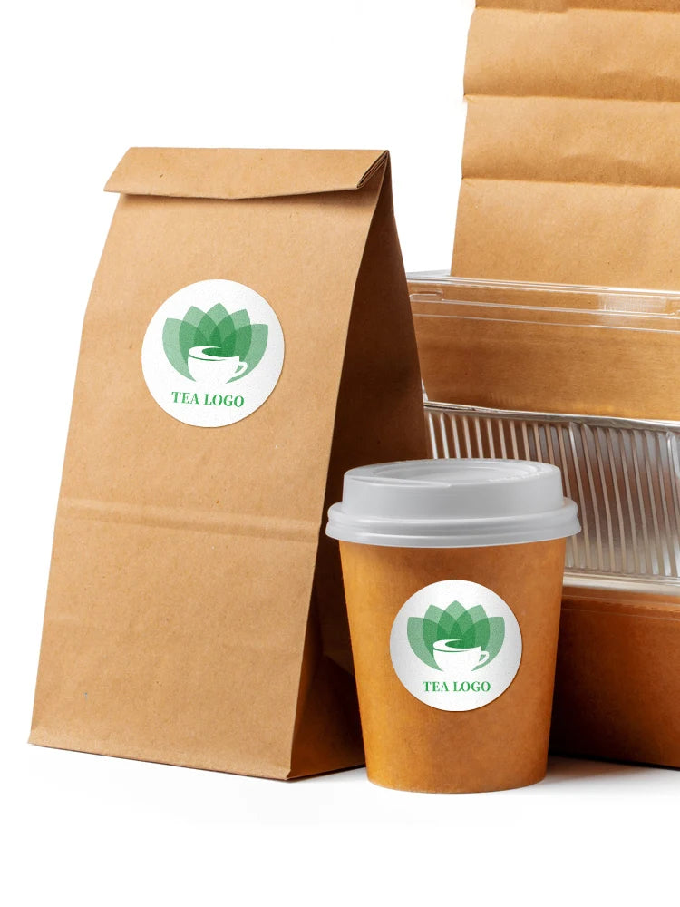 A brown paper bag and a disposable coffee cup both feature a green tea logo sticker.