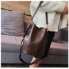 A brown alligator pattern crossbody bag with a leather strap is displayed next to a person sitting on a gray sofa.