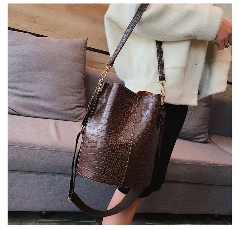 A brown alligator pattern crossbody bag with a leather strap is displayed next to a person sitting on a gray sofa.