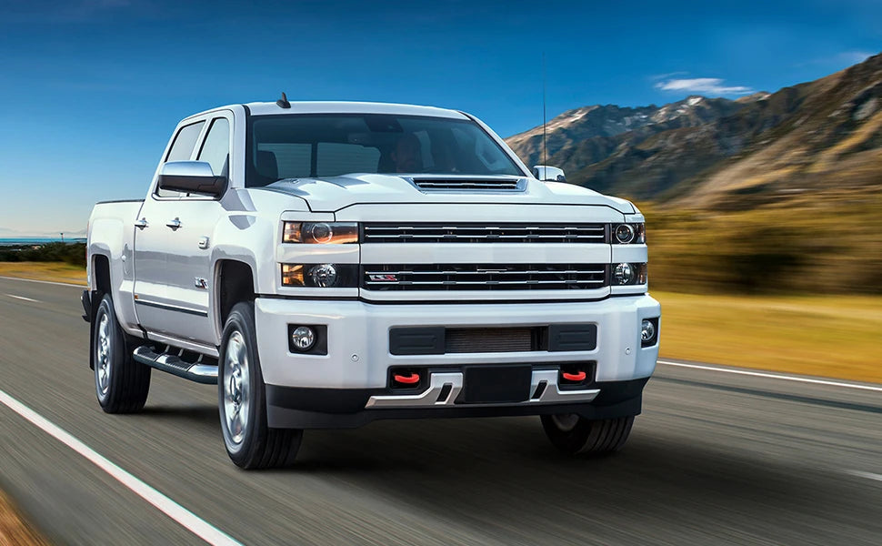A white Chevy Silverado pickup truck driving on a road with mountains in the background.