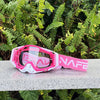 Pink and white HD Motocross goggles with anti-fog lens resting on a gray surface amidst green foliage.