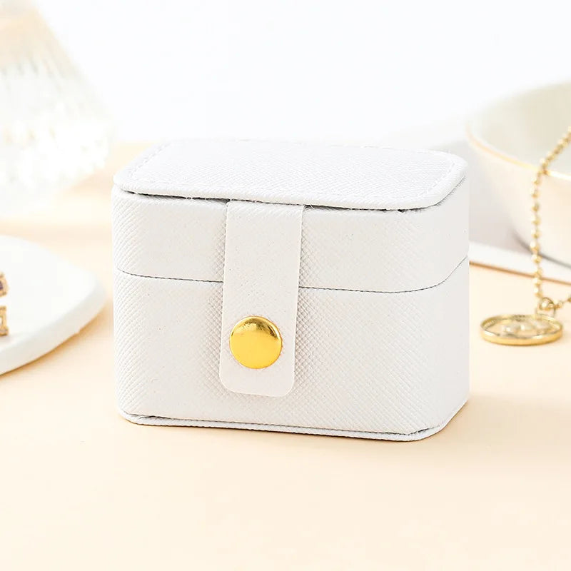 A white compact leather jewelry travel case with a gold-colored strap and closure is shown on a light surface.