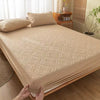 A beige quilted mattress protector is shown on a bed, with pillows and wooden furniture in the background.