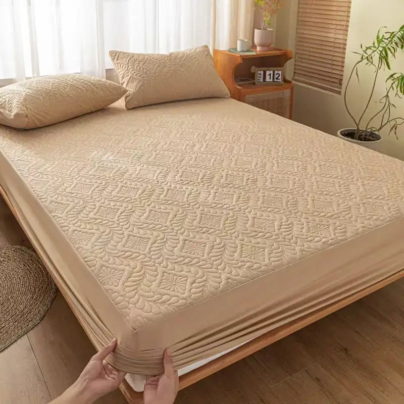 A beige quilted mattress protector is shown on a bed, with pillows and wooden furniture in the background.