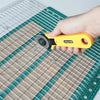 A hand holds a yellow acrylic quilting ruler with a dual scale grid and seam guide on a green cutting mat.