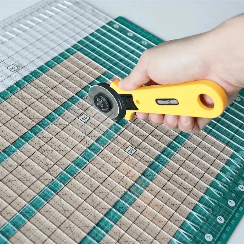 A hand holds a yellow acrylic quilting ruler with a dual scale grid and seam guide on a green cutting mat.