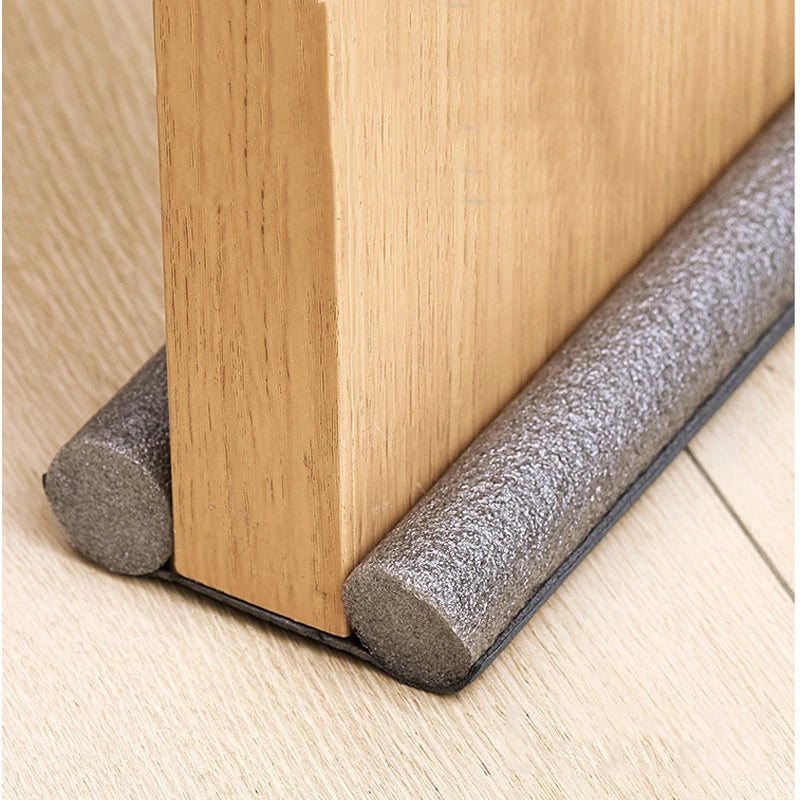 A close-up view of a wooden door frame with a gray flexible seal strip installed on the bottom edge.