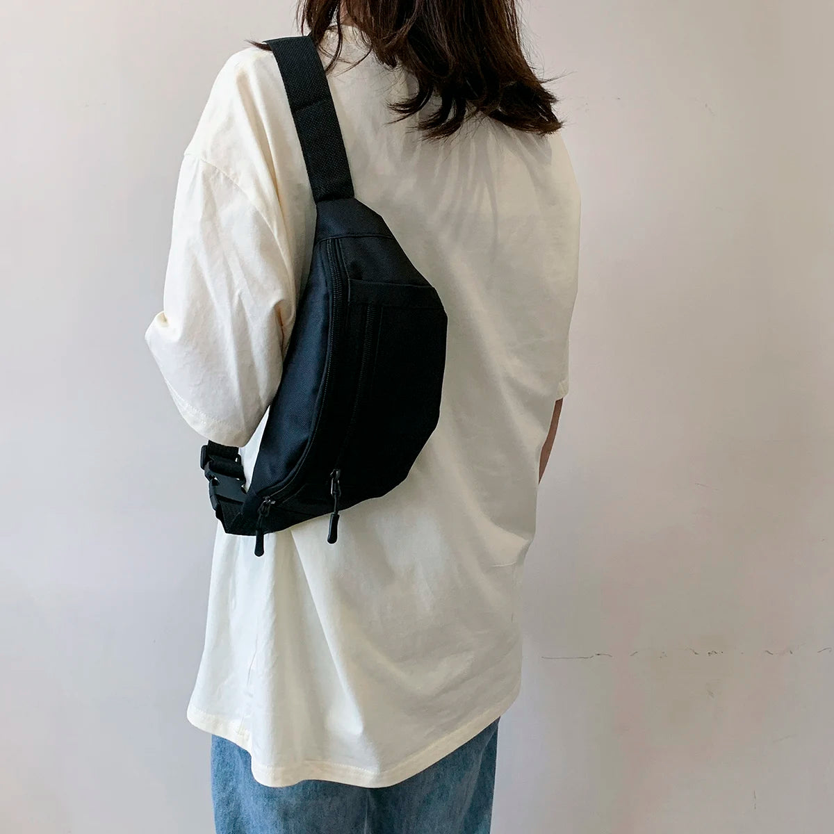 A person wearing a white t-shirt carries a black nylon crossbody waist pack on their back.