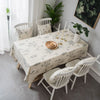 A dining table set with a cream tablecloth featuring a rustic fern print, surrounded by white wooden chairs.